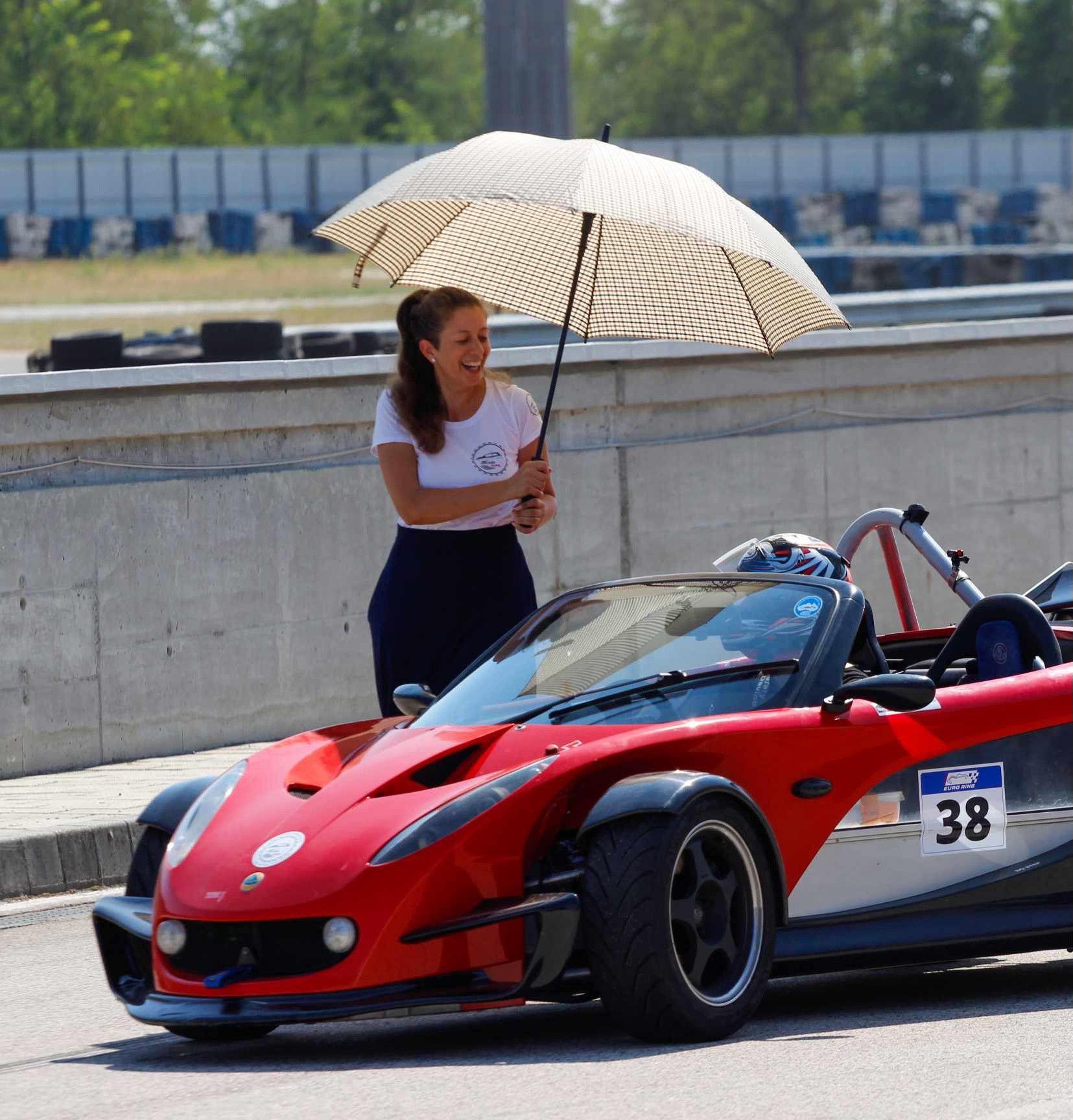 Piros Lotus 340R a bokszfalnál, mellette egy nő napernyővel